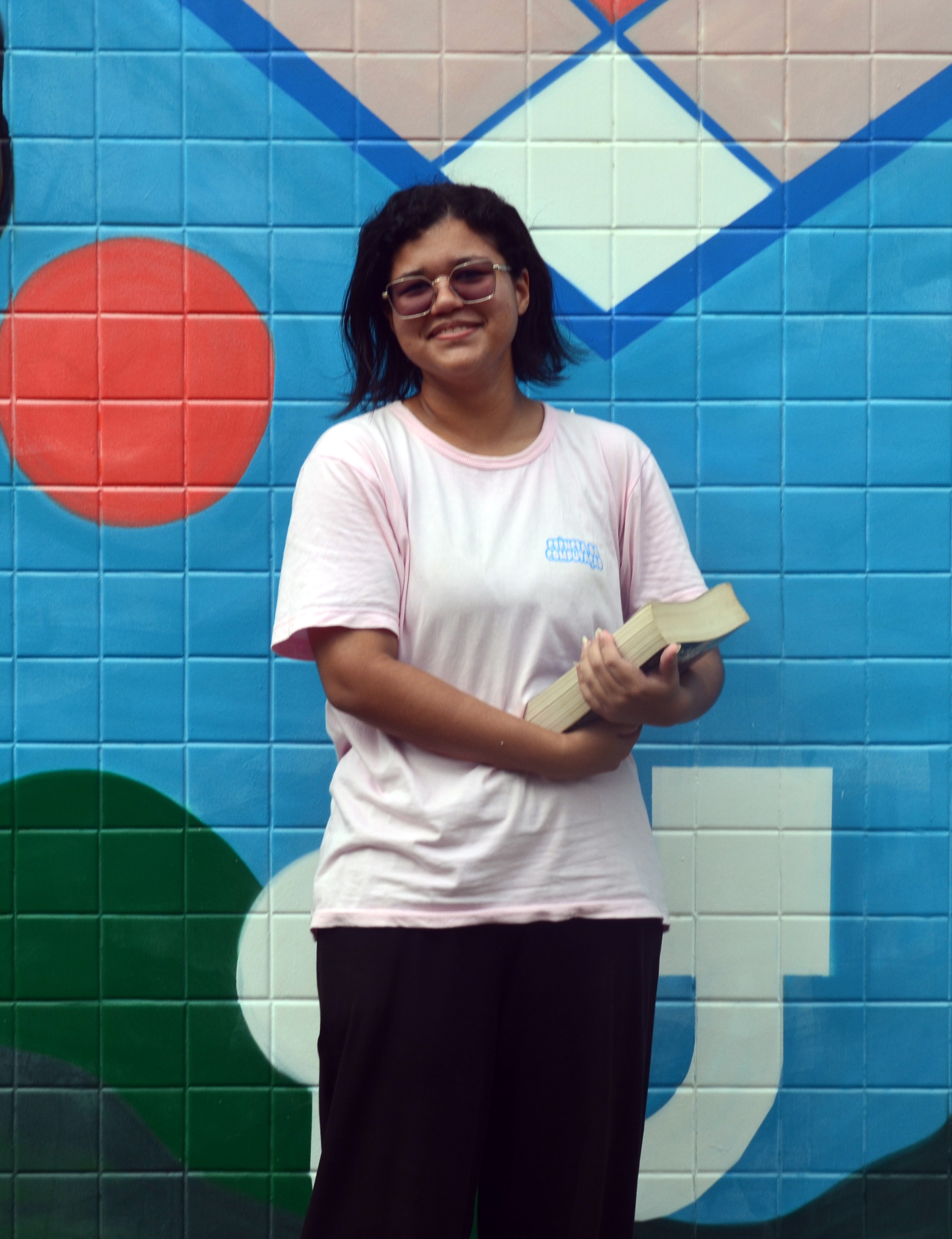 Mariana Santos, de blusa branca e calça preta  sorrindo segurando um livro
